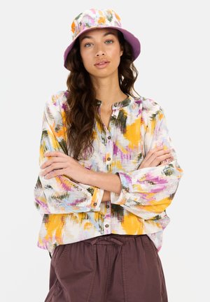 Kolorowa, bluzka z abstrakcyjnym wzorem, z długimi rękawami i guzikami, połączona z lawendowym kapeluszem typu bucket. Outfit charakteryzuje się miękkim, zwiewnym materiałem.