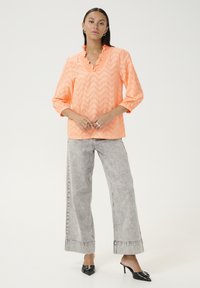 Blouse orange à motif zigzag texturé avec un col froncé et des manches 3/4, associé à un jean large gris et des talons aiguilles noirs.