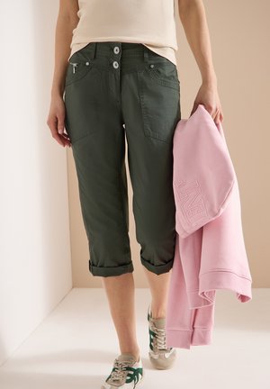 Personne portant un pantalon court retroussé vert foncé et des baskets beiges avec des détails verts, tenant un sweat-shirt rose dans la main droite.