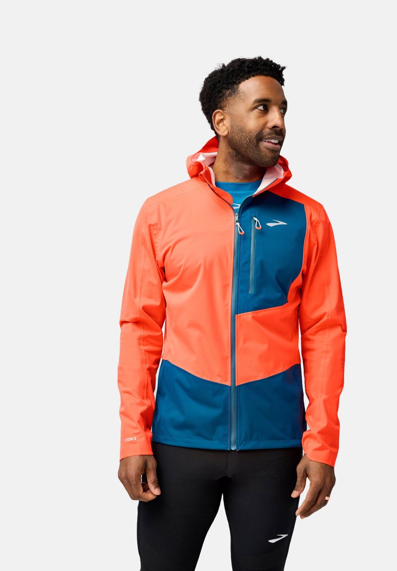 Veste pour hommes arborant un design en blocs de couleurs orange vif et bleu marine, fabriquée à partir d'un matériau léger et résistant à l'eau, dotée d'une capuche.