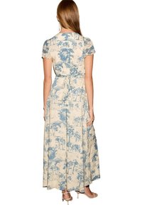 Femme aux cheveux longs portant une robe maxi beige avec des imprimés d'arbres bleus, manches courtes, et une ceinture nouée à la taille, debout, dos tourné vers l'avant.