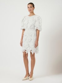 Γυναίκα με λευκό δαντελωτό φόρεμα με φουσκωτά μανίκια και λουλουδάτα σχέδια, συνδυασμένο με μπεζ ψηλοτάκουνα παπούτσια σε σχήμα αιχμής, που στέκεται μπροστά σε απλό φόντο.