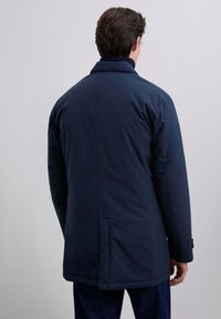Hombre vestido con un abrigo oscuro navy de longitud media y pantalones oscuros, de pie frente a un fondo liso gris claro con la espalda hacia el espectador.