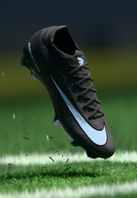 Black soccer cleat with textured synthetic material, featuring a prominent white Nike swoosh, situated above green grass with turf debris.