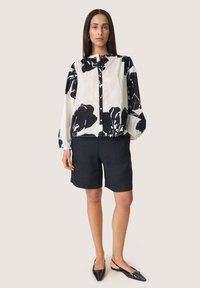 Weiße Bluse mit schwarzem Blumenmuster, knöpfbare Vorderseite, Ballonärmel; kombiniert mit dunklen Shorts und schwarzen spitzen Ballerinas.