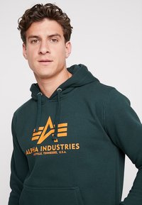 Felpa con cappuccio verde scuro, dotata di tasca a marsupio frontale. Presenta un logo arancione con "Alpha Industries" e un design geometrico. Coulisse regolabili.