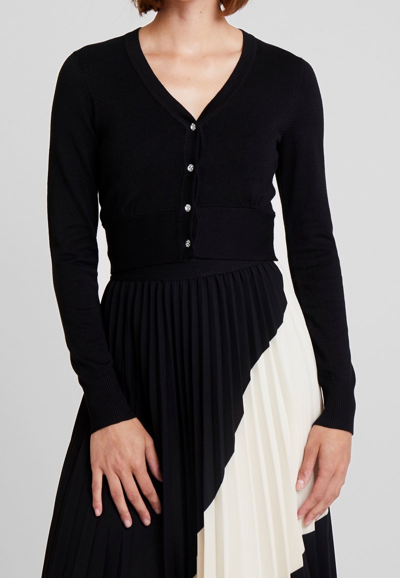 Femme portant un cardigan court noir avec des boutons associé à une jupe plissée asymétrique noire et blanche sur un fond uni.