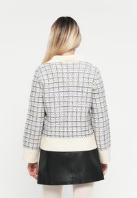 Vrouw met lang blond haar, gekleed in een wit en blauw geblokt gebreid sweater en een zwarte leren rok, staand met haar rug naar de kijker.