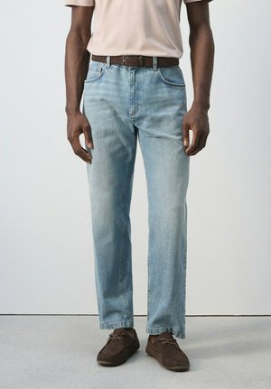 Uomo con jeans di denim azzurro chiaro, cintura marrone, t-shirt beige e scarpe stringate marrone scuro, in piedi davanti a uno sfondo semplice.