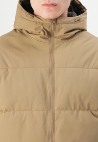 Padded jacket in a beige color, featuring a high collar, front zipper closure, and a textured surface with subtle vertical stripes.