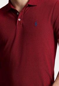 Czerwona koszulka polo z teksturowanej tkaniny, z klasycznym kołnierzykiem, z małym niebieskim logo na lewej piersi i białymi guzikami.