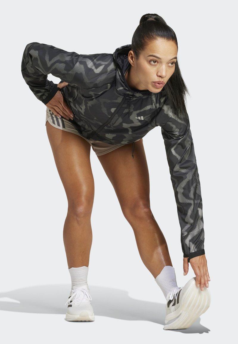 Leichte Jacke in Schwarz und Grau mit Muster und Kapuze. Model in Bewegung, trägt beigefarbene Shorts und weiße Sneakers mit grauen Akzenten.