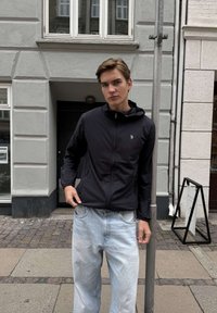 Noor mees musta lukuga jope ja helelillas teksapükstes seisab linna tänaval metallposti ja hoone sissepääsu kõrval.