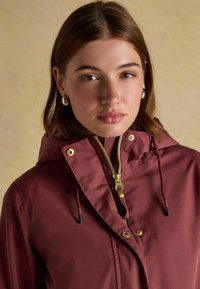 Burgundowa kurtka z kapturem z złotym zamkiem, sznurkami z czarnymi końcówkami i złotymi napami. Tkanina wydaje się gładka, o minimalnej teksturze.