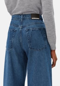 Jeans en denim bleu taille haute avec une coupe droite, de grandes poches et une étiquette de marque noire à l'arrière de la taille. Texture lisse.