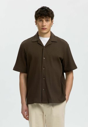 Jeune homme portant une chemise marron côtelée à manches courtes boutonnée sur un sous-vêtement blanc et un pantalon beige, debout devant un fond uni.