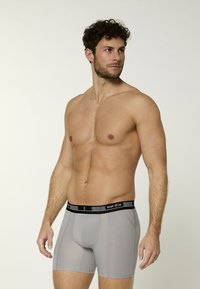 Un modello maschile si trova lateralmente indossando boxer grigi con cintura nera, in uno sfondo neutro e con un'espressione sicura.