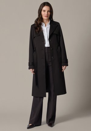 Kobieta w długim czarnym płaszczu trencz, na białej koszuli i czarnych szerokich spodniach, stojąca na jednolitym jasnoszarym tle.