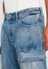 Világoskék denim munkanadrág, kopott hatással, több zsebbel és elöl gombos záródással. Lekerekített szegély, normál varrással.