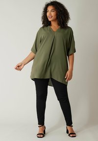 Olivgrünes, kurzärmliges Oberteil mit V-Ausschnitt und plissiertem Design. Kombiniert mit schwarzen Skinny-Jeans und schwarzen, trägerlosen Schuhen.