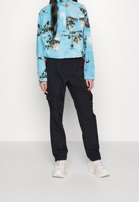 Person wearing a blue floral and bird-patterned jacket, black cargo pants, and white chunky sneakers standing against a plain white background.