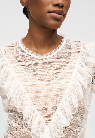 Gros plan sur une personne portant un chemisier en dentelle blanche avec des détails volantés et des motifs floraux, montrant le décolleté et le haut du torse.