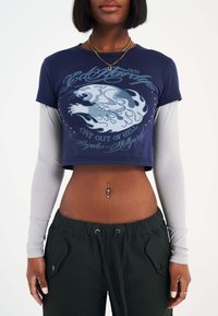 T-shirt cropped blu navy con design grafico di un leone, sovrapposto a una maglietta a maniche lunghe grigia chiara; pantaloni neri con cintura elastica.