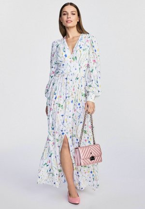 Kwiatowa sukienka maxi z długimi rękawami i głębokim dekoltem w kształcie litery V, z bocznym rozcięciem. Biała tkanina z wielokolorowymi wzorami kwiatowymi. Różowa torebka.