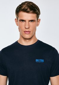 Noor mees lühikeste pruunide juustega, kes kannab tumesinist T-särki, millel on rinnal sinise tekstiga "BIG STAR", seisab valgusküllase tausta ees ja vaatab otse ette.