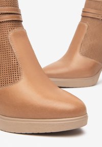 Bottines à talons en cuir lisse de couleur beige, avec des détails perforés et un semelle en coin. Bout arrondi et accents de couture discrets.