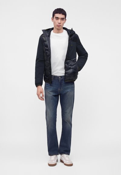 Jeune homme portant une veste matelassée noire sur un t-shirt blanc, un jean bleu et des baskets blanches, debout devant un fond uni.