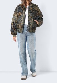 Zielono-czarna bomberka z nadrukiem o luźnym kroju, z prążkowanymi mankietami i dużymi kieszeniami, w zestawie z jasnoniebieskimi workowatymi dżinsami i białymi sneakersami.