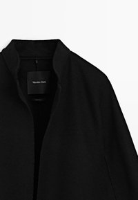 Cappotto nero aperto davanti con colletto alto, che mostra l'etichetta del marchio Massimo Dutti all'interno del collo su uno sfondo bianco.