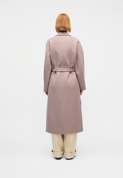 Άτομο με κοντά κόκκινα μαλλιά που φοράει ένα μωβ παλτό με ζώνη και μπεζ παντελόνι με φαρδιά πόδια, στέκεται πλάτη σε μονόχρωμο λευκό φόντο.