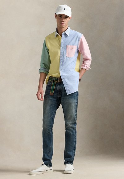 Ung mand iført en flerfarvet skjorte med knapper, blå jeans, hvide sneakers, hvid kasket og stribet bælte, står mod en ensfarvet baggrund.