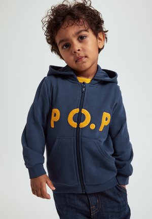 Ungt barn med krøllet hår iført en mørkeblå hættetrøje med lynlås og gule "P.O.P" bogstaver, stående foran en ensfarvet baggrund.