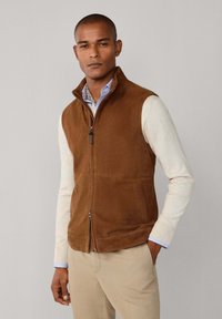 Braune Wildlederweste mit hohem Kragen, Reißverschlussverschluss und Seitentaschen. Über einem leichten Pullover und einem gemusterten Hemd getragen, was für eine mehrschichtige Textur sorgt.