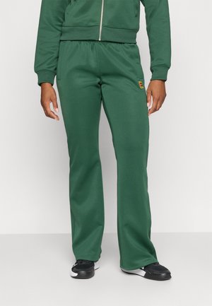 Personne portant une veste zippée verte et un pantalon de jogging vert assorti avec des baskets noires et blanches, visible de la taille aux pieds.