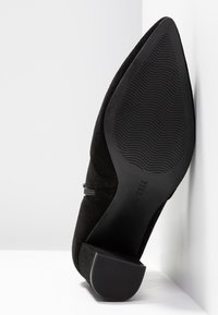 Zapato de tacón alto de ante negro con puntera puntiaguda y suela de goma texturizada, que presenta una sutil correa de acento en el lateral.