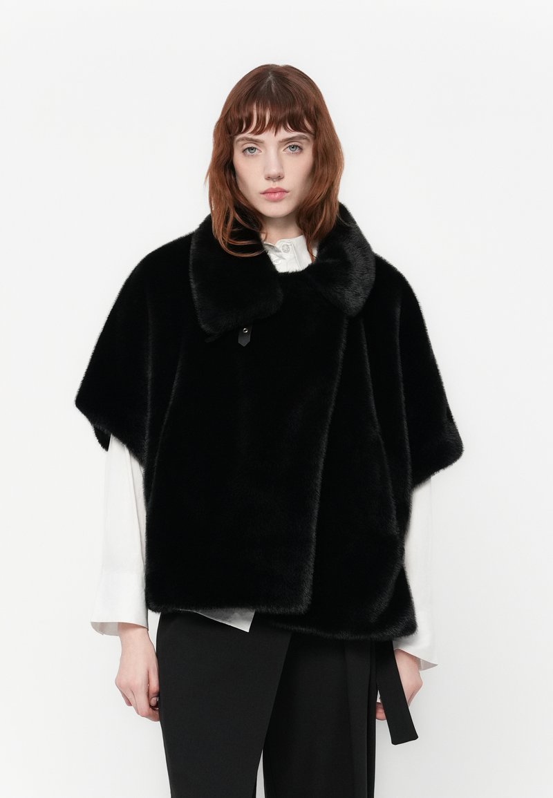 Vrouw met rood haar, gekleed in een zwart nepbont kort cape over een witte blouse en zwarte broek tegen een witte achtergrond.