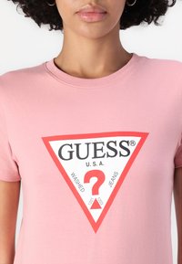 Gaiši rozā kokvilnas T-krekls ar sarkanu trīsstūra logotipu, kurā ir teksts "GUESS®", "WASHED" un "JEANS". Īsas piedurknes un apaļa kakla izgriezums.