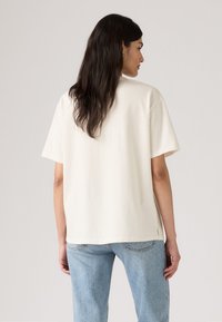 T-shirt en coton couleur crème avec une coupe décontractée, des manches courtes et des fentes sur les côtés. Associé à un jean en denim bleu clair, légèrement usé.