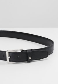Cinturón de cuero negro con acabado texturizado, hebilla rectangular de plata y cierre de un solo pasador. Acertado con un logotipo en el lazo del cinturón.