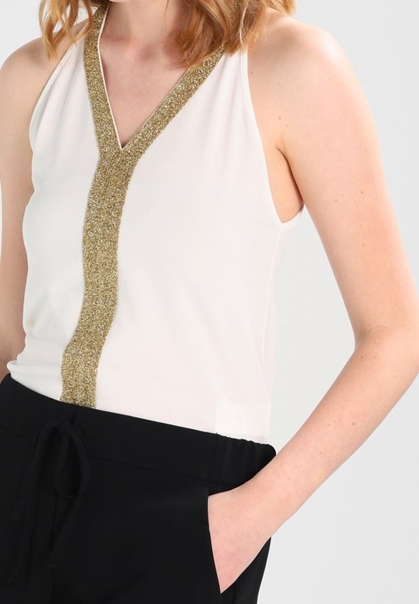 Femme portant un haut blanc sans manches avec une bordure en V pailletée dorée, associé à un pantalon noir, main dans la poche, sur un fond uni.