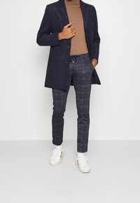 Marineblazer, bruine coltrui, grijs geblokt broek met koord, en witte sneakers. De blazer heeft twee knopen en een getailleerde pasvorm.