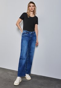 Černé krátké rukávy tričko v kombinaci s širokými modrými džíny z denimu. Džíny mají klasický střih s viditelným šitím a kapsami. Bílé tenisky.