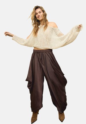 Femme souriante aux cheveux ondulés détachés, portant un pull en maille crème à épaules dénudées, un pantalon large marron et des bottes pointues marron.