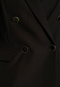 Blazer negro con una textura suave, que presenta dos botones negros con borde dorado y un diseño de solapa cl�ásico.