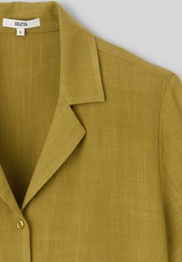 Blouse vert olive en tissu texturé, dotée d'un col à revers et d'une fermeture à un bouton. Étiquette de taille "S" visible.