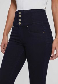 Tmavě modré džíny s vysokým pasem, které mají široký pas s pěti zlatými knoflíky, přední kapsy a decentní švy.